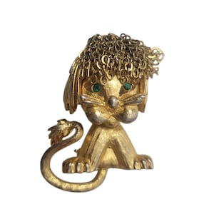 May include: A gold-toned lion brooch with green rhinestone eyes and a chain mane. The lion is in a seated position with a curled tail. The brooch has a textured finish and is set against a white background.
