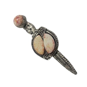 May include: A silver-toned brooch in a dagger shape. The brooch features two oval-shaped, cream-colored stones with red speckles. The handle and blade are detailed with textured patterns and a rounded, speckled stone at the top.