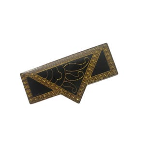 May include: A rectangular, Art Deco-style brooch with a black enamel center and gold-toned metal trim. The brooch features geometric shapes and a stylized sunburst design. The edges are adorned with small, circular details.