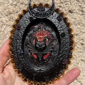 Peut inclure: Un cadre décoratif noir de forme ovale avec un motif de chauve-souris et de croissant de lune. Le cadre a une bordure perlée dorée et noire et de petits accents de roses. La chauve-souris a des accents rouges.