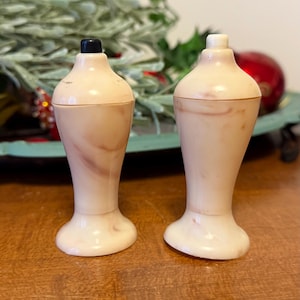 May include: Two vintage salt and pepper shakers. The shakers are a light pink color with a marbled effect. One has a black top, and the other has a white top. The shakers have a flared base and a narrow neck.