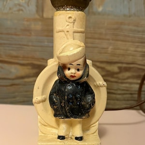 May include: Vintage sailor-themed lamp with a cream-colored base and a decorative sailor figurine. The sailor wears a navy blue coat and a white hat. The lamp has a brown metal top and a cord.
