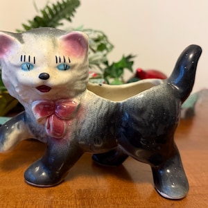 May include: A ceramic cat planter with a gray and white face, blue eyes, and a pink bow. The cat has a black tail and legs. The planter is designed to hold small plants or flowers.