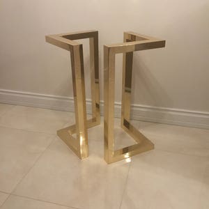 May include: Gold metal table base with a geometric design. The base is made of two square frames that are connected at the top and bottom.