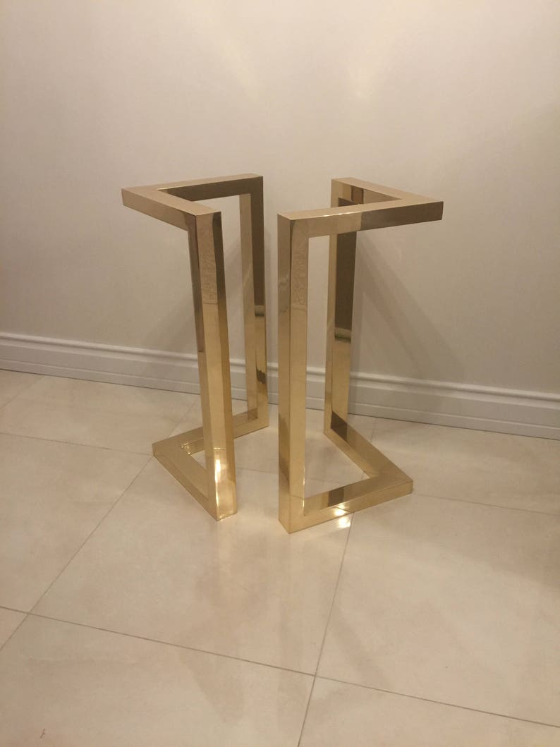 May include: Gold metal table base with a geometric design. The base is made of two square frames that are connected at the top and bottom.