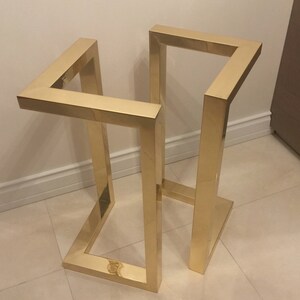 May include: A gold metal stand with a geometric design. The stand is made of square tubing and has a rectangular base.