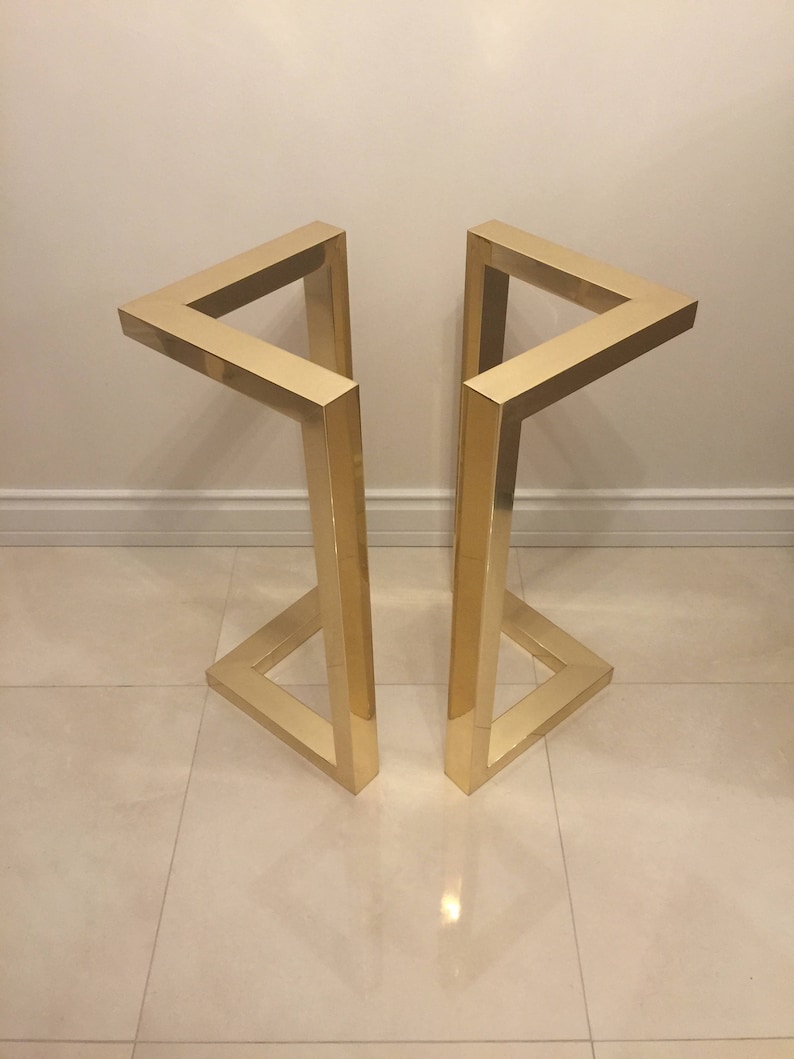 May include: Two gold metal table legs with a geometric design. The legs are made of square tubing and are connected at the top and bottom to form a triangular shape.