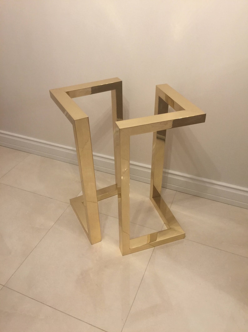 May include: A gold metal stand with a square base and four legs. The stand is made of a single piece of metal that is bent into a square shape.