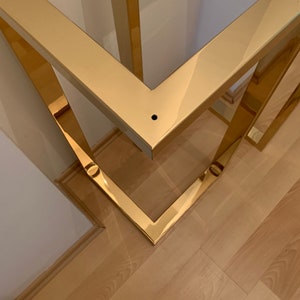 May include: Close-up of a gold metal table base with a square design. The base is made of polished metal and has a shiny finish.