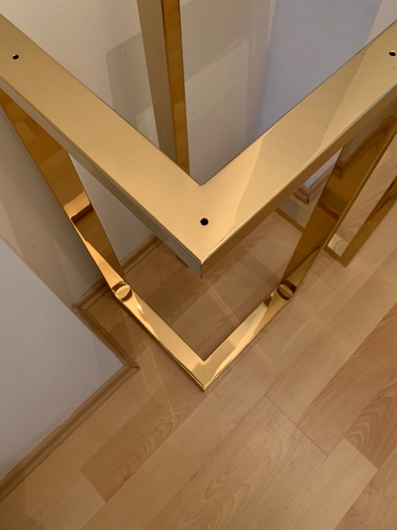 May include: Close-up of a gold metal table base with a square design. The base is made of polished metal and has a shiny finish.