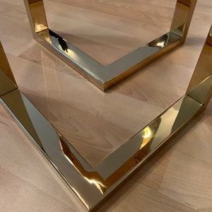 May include: Two gold metal table legs form a V-shape. The legs are polished and reflect the surrounding light.