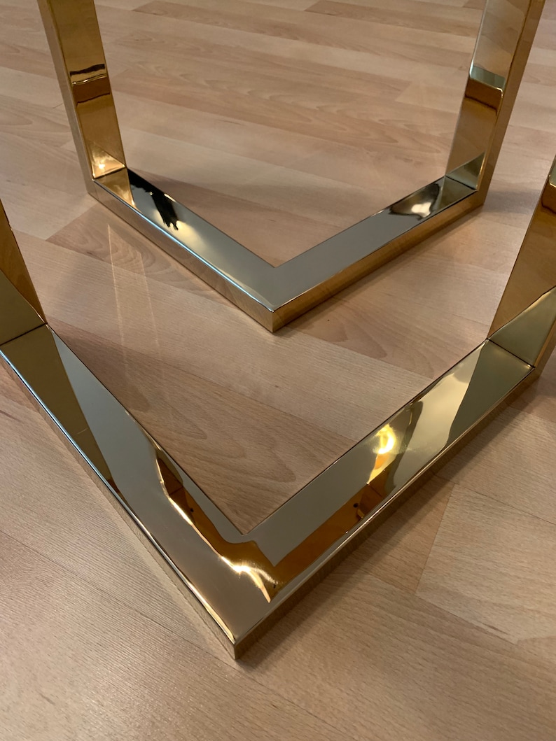 May include: Two gold metal table legs form a V-shape. The legs are polished and reflect the surrounding light.