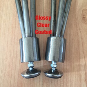 May include: Close-up of two metal table legs with a glossy clear coated finish. Each leg has a threaded metal base with a black rubber foot.