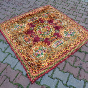 Puede incluir: Una alfombra vintage de forma cuadrada con un patrón floral en tonos rojos, amarillos y verdes. La alfombra tiene un borde con flecos.