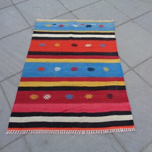 May include: A colorful striped rug with a geometric pattern. The rug has red, yellow, blue, black, and white stripes with small, colorful geometric shapes woven into the design.