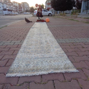 Puede incluir: Una alfombra rectangular de estilo vintage, con un fondo crema y patrones intrincados en azul y beige. La alfombra está colocada sobre una superficie pavimentada con ladrillos, con una escoba y una figura decorativa cerca.