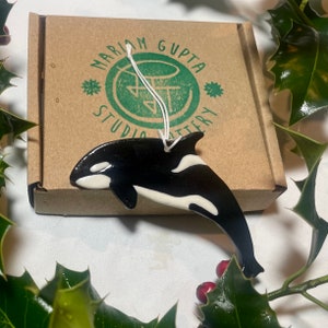 May include: A white and black ceramic ornament shaped like an orca whale. The ornament is hanging from a white string and is packaged in a brown box with a green logo that reads "Mariam Gupta Studio Pottery".