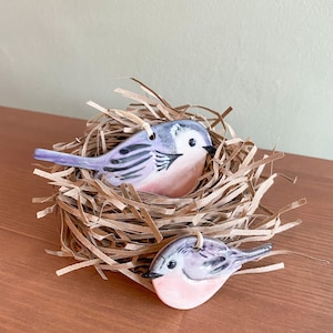May include: Two ceramic bird ornaments in a nest made of brown paper strips. The birds have lavender and pink colouring with black details. The nest sits on a brown wooden surface.