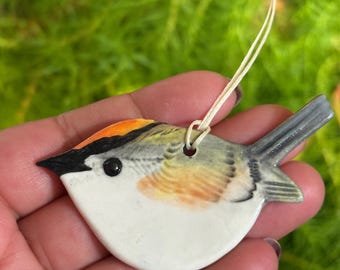 May include: A ceramic bird ornament with a white belly, orange head, and gray tail. The bird has a black eye and beak. A cream-colored string is attached for hanging. The ornament is designed to look like a real bird.