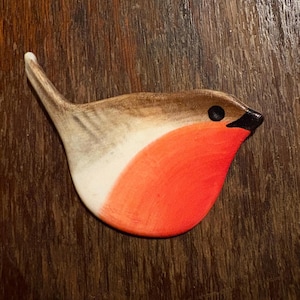 May include: A wooden robin ornament with a brown and white head, a vibrant red breast, and a black beak. The bird is set on a wooden surface. The ornament is a decorative item.