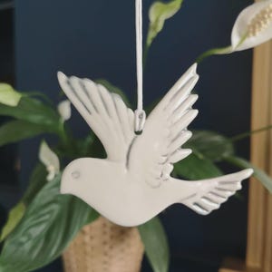 May include: A white ceramic dove ornament with gray detailing on the wings, hanging from a white string. The dove is in flight, with its wings spread. The background includes green plants and a woven basket.