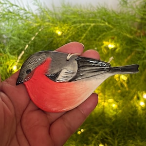 May include: A ceramic bird ornament with a red breast, grey head, and black tail. The bird is held in a hand against a backdrop of green foliage and small, warm lights. The ornament has a string for hanging.