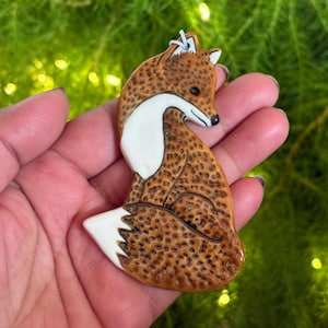 Puede incluir: Figura de zorro de cerámica con acabado brillante. El zorro es principalmente marrón con un patrón moteado, un pecho blanco y una cola con punta blanca. El zorro está en posición sentada, con la cabeza girada hacia un lado.