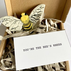 May include: A ceramic bee figurine with black and yellow stripes and white wings, packaged in a brown box with white tissue paper. The box also includes a white card with the text "You're the bee's knees."