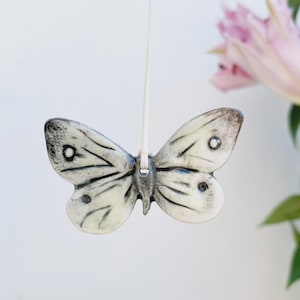 May include: A ceramic butterfly ornament with a cream-coloured body and black wing details. The butterfly has a string attached for hanging. The background is a soft white with a blurred pink flower.