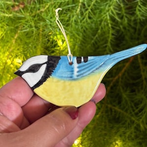 May include: A ceramic bird ornament with a yellow belly, blue wings and tail, and a white head with black accents. The bird has a string for hanging. The ornament is held in a hand against a green background.