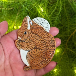 May include: A ceramic squirrel ornament with a brown body, white belly, and a white and dotted tail. The squirrel is holding something in its paws. The ornament has a loop for hanging.
