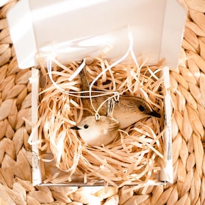 May include: Two bird ornaments in a white box filled with shredded paper. The ornaments are light brown with black eyes and are hanging by white strings. The box is on a woven, light brown surface.