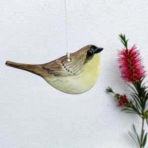 May include: A hand-painted ceramic bird decoration. The bird has a brown back, a yellow breast, and a black beak. It hangs from a white string. A red bottlebrush flower is visible in the background.