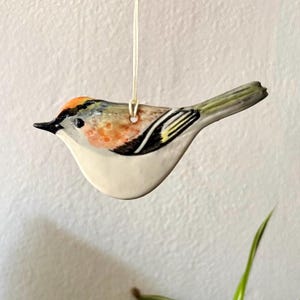 Könnte beinhalten: Ein Keramik-Vogelornament mit cremefarbenem Bauch, schwarz-weißem Flügelmuster und einem orangefarbenen Fleck auf dem Kopf. Der Vogel hängt an einer dünnen weißen Schnur, die am Rücken befestigt ist, und hat eine glänzende Oberfläche mit Bemalung.