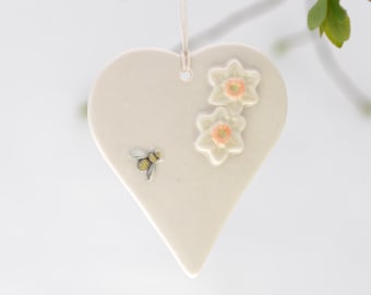 May include: White ceramic heart-shaped ornament with two white daffodils and a bee. The daffodils have orange centers.