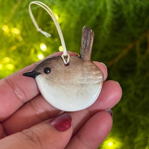 May include: A ceramic bird ornament with a white belly, brown back, and black beak. The bird has a small, brown tail and a cream-coloured string for hanging. The ornament is held in a hand against a blurred green background.