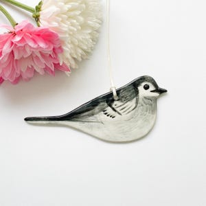 May include: A ceramic bird ornament in shades of grey and white, suspended by a white string. The bird has a detailed painted design. Pink and white flowers are in the upper left corner, set against a white background.