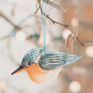 May include: A ceramic bird ornament with a blue and orange design, suspended by a light blue string. The bird is on a tree branch with a blurred background. The ornament is approximately 10 cm long.
