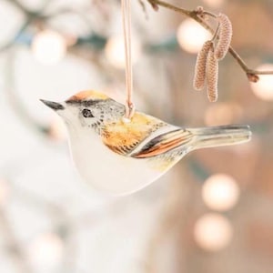 May include: A ceramic bird ornament with a white body, orange and brown wings, and a tan head. The bird is suspended from a thin, pale pink string. The background is blurred with soft, warm tones.