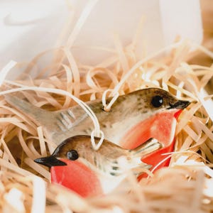 May include: Two ceramic robin ornaments with red breasts, brown wings, and black beaks sit in a bed of straw. Each bird has a white string for hanging. The ornaments are detailed and appear handmade.