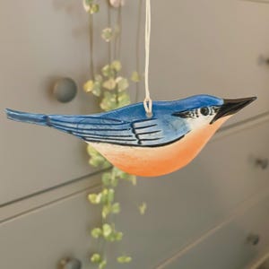 May include: A ceramic bird ornament with a blue back and wings, an orange breast, and a black beak. The bird is suspended by a white string. The ornament is a decorative item.