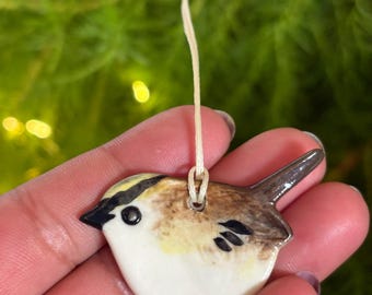May include: A ceramic bird ornament with a white belly, brown and yellow head, and a dark brown tail. The bird has a black eye and is suspended by a thin, off-white string. The ornament is held in a person's hand.