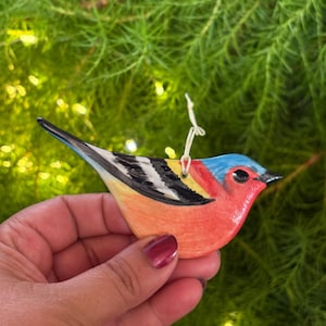 Könnte beinhalten: Ein Keramik-Vogelornament mit einem lebendigen Farbschema. Der Vogel hat eine rote Brust, einen blauen Kopf und schwarz-weiße Flügel. Zum Aufhängen ist eine weiße Schnur angebracht. Das Ornament wird vor einem Hintergrund aus grünem Laub gehalten.