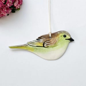 Puede incluir: Adorno de pájaro de cerámica con vientre color crema, cuerpo amarillo-verdoso y alas marrones y negras. El pájaro tiene un pico negro y está suspendido por una cuerda blanca. Flores rosas en el fondo.