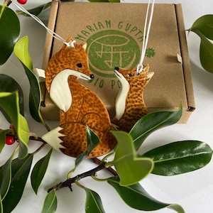 May include: Two ceramic fox ornaments, one larger than the other, with brown and white paint. The foxes are facing each other and have a sweet expression. The ornaments are hanging from white string and are in front of a brown box with the text "MARIAM GUPTA" printed on it.
