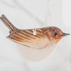 May include: A ceramic bird ornament featuring a glossy finish with brown and tan plumage details on the wings and head, a cream-coloured belly, and a dark eye. A small hole is located on the back for hanging with a thin white string.