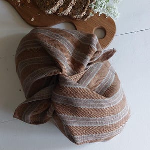 May include: A loaf of seeded bread, partially sliced, rests on a wooden cutting board. A brown and tan striped cloth is knotted at the bottom. White flowers are arranged on the right side of the image. The bread appears to be homemade.