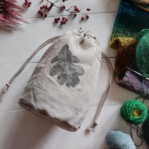Op de afbeelding: Een beige tas met trekkoord van geweven stof met een donkergrijs bedrukt eikenbladontwerp op de voorzijde. De tas heeft twee bijpassende beige koorden aan de bovenkant en is geconstrueerd met zichtbare verticale en horizontale naden.