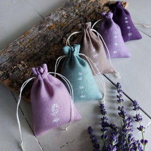 Peut inclure: Cinq petits sachets en tissu avec cordon de serrage dans des tons lavande, bleu sarcelle, beige, violet clair et violet foncé. Chaque sachet présente un motif de brin botanique imprimé en blanc et est resserré par un cordon blanc.