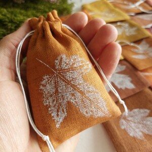 May include: A small, rectangular, ochre-coloured linen drawstring pouch featuring a white screen-printed maple leaf design on the front. The pouch is secured with thin white cord strings that are knotted at the ends, creating a gathered top closure.